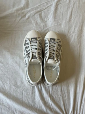 Vita sneakers från Dior med monogram - Säljer ett par vita sneakers från Dior med tydligt monogrammönster i grått och svart. Skorna har snörning, rund tå och en unik stjärndetalj under sulan. Klassisk och trendig design med Dior-logga på plösen.