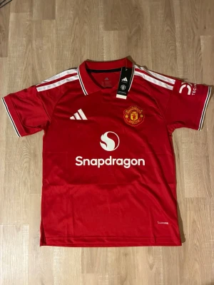 Manchester United hemmatröja 26/27 - Snygg röd Manchester United matchtröja från säsongen 26/27 med vita detaljer och klubbmärke på bröstet. Tröjan har tryck med 'Snapdragon' på framsidan och 'B. Fernandes 8' på ryggen. Tillverkad i lätt och ventilerande material, perfekt för fotboll eller träning. HELT NY!