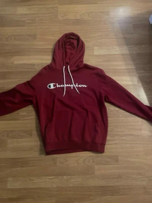 Röd Champion hoodie med huva - Säljer en snygg röd hoodie från Champion med vit logga på bröstet och vita dragsnören i huvan. Hoodien har en stor magficka och är gjord i mjukt bomullsmaterial. Perfekt för en avslappnad och sportig stil. Testade den två gånger så den är i princip ny.
