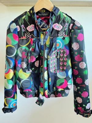 Färgglad bomberjacka från Desigual strl S - Unik bomberjacka från Desigual med färgglada cirklar och blommor i rosa, blått, grönt och gult på svart botten. Jackan har dragkedja, fickor framtill och broderade detaljer. Perfekt för dig som vill sticka ut med din stil.