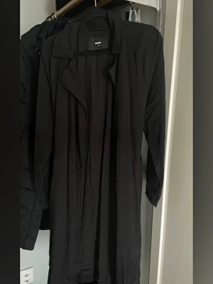 Svart trenchcoat med klassisk krage - Stilren svart trenchcoat med klassisk krage och långa ärmar. Modellen har öppen front utan synliga knappar och ett mjukt jeans material som ger ett snyggt fall. Perfekt för lager-på-lager och en tidlös look.