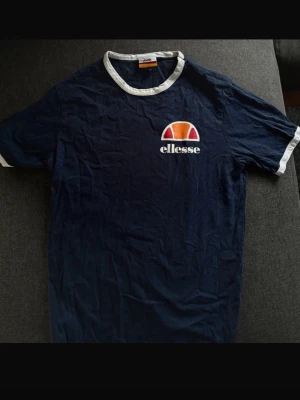 Marinblå T-shirt från Ellesse - Marinblå tröja av märket Ellesse. - vit krage samt vid ärmsluten. - lång form - normal passform med viss rymlighet  - mycket bra skick men med mindre sprickor i trycket framtill (se bild 3).  Storlek: XS men passar även