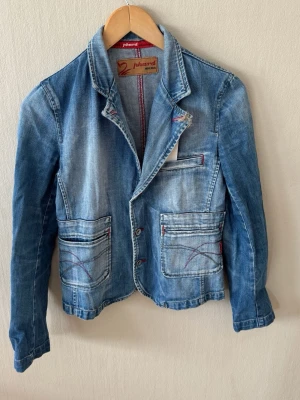 Phard Blå jeansjacka med kavajkrage - Snygg blå jeansjacka från Phard med kavajkrage och två stora fickor framtill. Jackan har röda sömdetaljer och knappar vid ärmsluten. Unik design som sticker ut med sin tvättade denimlook och avslappnade passform. 0075