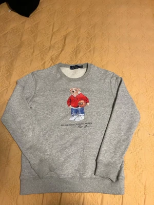 Grå sweatshirt Polo Bear Ralph Lauren - Grå sweatshirt från Ralph Lauren med det klassiska Polo Bear-motivet på bröstet. Tröjan har rund hals, ribbade muddar och är tillverkad i mjukt material. Perfekt för en avslappnad och trendig look.