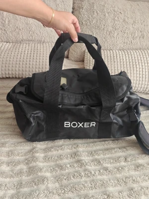 Svart väska BOXER - Svart väska med BOXER-tryck. Väskan har dubbla bärhandtag, justerbar axelrem och en praktisk ytterficka med dragkedja. Tillverkad i slitstarkt syntetmaterial med enkel, sportig design och plats för det mesta du behöver.