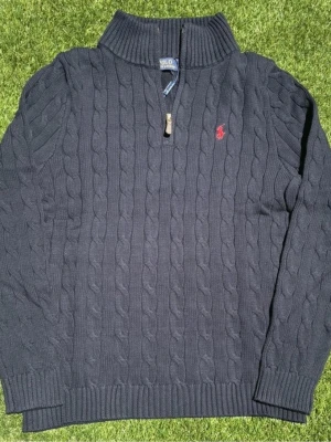 Mörkblå kabelstickad tröja Polo Ralph Lauren - Kabelstickad mörkblå tröja från Polo Ralph Lauren med halv dragkedja och hög krage. Tröjan har ett rött broderat logomärke på bröstet, ribbade muddar och nederkant. Perfekt för dig som gillar klassisk och stilren look.