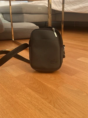 Svart axelväska från Lacoste ”Äkta” - Snygg svart axelväska från Lacoste med justerbar axelrem och diskret logga framtill. Väskan har en kompakt, rektangulär form och är tillverkad i slitstarkt material med dragkedja upptill och en extra ficka på sidan. Perfekt för att bära det viktigaste.