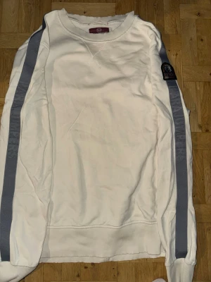 Vit sweatshirt med grå ränder - Vit sweatshirt med grå ränder längs ärmarna och en patch på ena ärmen. Tröjan har rund halsringning och ribbade muddar vid ärmslut och nederkant. Perfekt för en avslappnad och sportig stil.