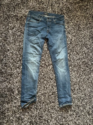 Blå slim fit jeans - Säljer ett par blå slim fit jeans med klassisk femficksdesign och lätt slitna detaljer framtill. Jeansen har normal midja och smala ben, perfekta till en avslappnad stil. Materialet är denim i bomull med lite stretch för extra komfort.