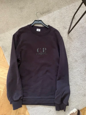 Svart sweatshirt från C.P. Company - Stilren svart sweatshirt från C.P. Company med rund halsringning och diskret logga på bröstet. Tröjan har ribbade muddar vid ärmslut och nederkant, perfekt för en avslappnad och trendig look.