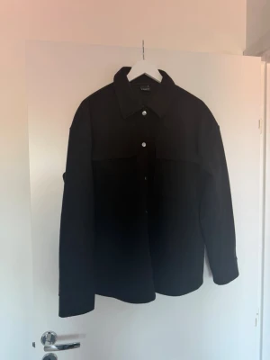 Svart overshirt med knappar - Stilren svart overshirt med klassisk krage och knäppning framtill. Overshirten har raka ärmar och två bröstfickor. Perfekt att slänga över en t-shirt för en avslappnad look. 