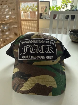   Chrome Hearts keps - Säljer en camouflagemönstrad truckerkeps från Chrome Hearts med svart mesh på sidorna och bak. Framtill sitter en stor svart patch med vit text: 'CHROME HEARTS FUCK HOLLYWOOD, USA'. Justerbar baktill och riktigt streetwear-vibe.