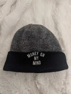 Gråmelerad mössa med text - Säljer en gråmelerad mössa med svart uppvikt kant där det står 'MONEY ON MY MIND' broderat i vitt. Mössan är stickad och har en klassisk form som passar perfekt till streetwear eller casual outfits.