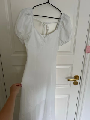Vit långklänning med puffärm - Säljer en elegant vit långklänning med puffärmar och knyt i ryggen. Klänningen har en feminin siluett och fyrkantig ringning. Perfekt för sommarens alla tillfällen. Materialet känns lätt och luftigt.
