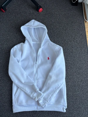 Vit hoodie med dragkedja och huva - Säljer en vit hoodie med dragkedja och huva. Tröjan har två fickor framtill och en liten röd logga broderad på bröstet. Klassisk och enkel design som passar till det mesta. Mjuk och skön bomullskänsla.