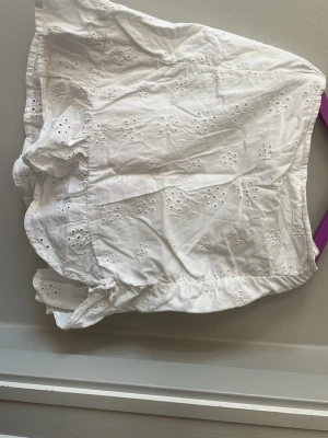 Vit kort kjol med broderade detaljer - Söt vit kort kjol med broderade blommönster och en liten rosett längst ner. Kjolen har volangkant och på bakre sidan där uppe har kjolen en stretchigt kant.