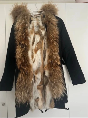 Päls jacka - Svart exklusiv vinterparkas med äkta päls. Jackan har ett tjockt och mjukt pälsfoder på insidan samt en stor, fyllig pälskant runt huvan och framtill i vackra bruna, beige och ljusa nyanser.  Väldigt varm och perfekt för höst och vinter. Snygg figurnära modell med lyxig look. Pälsen är tät och välbevarad.  ✨ Äkta päls ✨ Varmt pälsfoder ✨ Stor pälskant på huvan ✨ Svart yttertyg ✨ Mycket fint skick