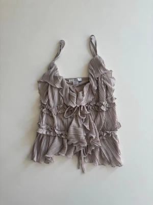 Beige omlottopp med volanger - Supersöt beige omlottopp med volangdetaljer och knyt framtill. Toppen har tunna axelband och ett plisserat tyg som ger en romantisk känsla. Perfekt för dig som gillar en feminin och trendig stil.