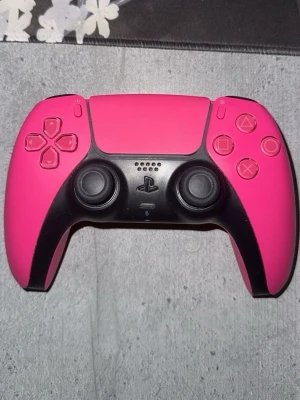 Rosa PlayStation-handkontroll - Snygg rosa handkontroll till PlayStation med svart detalj runt styrspakar och knappar. Ergonomisk form med klassiska PlayStation-symboler på knapparna. Tillverkad i plast och passar perfekt för gaming.