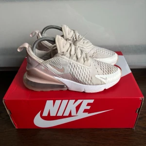 Nike Air Max 270 beige sneakers - Snygga Nike Air Max 270 sneakers i beige och vitt med mesh-överdel och stora synliga Air Max-dämpning i hälen. Skorna har vita snören, diskreta loggor och en modern, sportig siluett. Perfekta för dig som gillar streetwear och komfort.