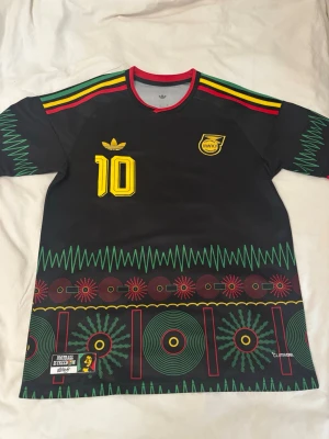 Jamaica Marley fotbollströja svart - Snygg svart fotbollströja med färgglada detaljer i rött, gult och grönt. Nummer 10 och Marley tryckt i gult, samt Jamaica-märke på bröstet. Unik grafisk design längs nederkant och ärmar. Perfekt för dig som gillar reggae och sportig stil.
