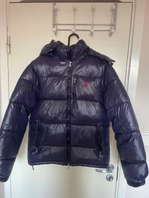 Ralph Lauren Mörklila jacka - Snygg mörklila pufferjacka med avtagbar huva och dragkedja framtill. Jackan har två sidofickor med dragkedja och en bröstficka med liten röd logga. Perfekt för kalla dagar och håller dig varm hela vintern.