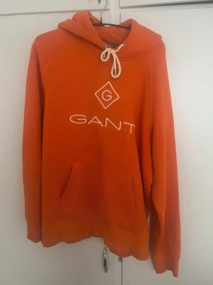 Orange hoodie från GANT med logga - Snygg orange hoodie från GANT med stor vit logga och G-symbol på bröstet. Hoodien har huva med vita dragsnören och en stor magficka framtill. Perfekt för en chill och avslappnad stil. Har änvadt några gånger 