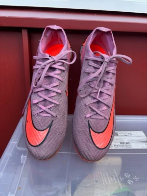 Lila Nike elite fotbollsskor med orangea detaljer  - Säljer ett par snygga lila Nike fotbollsskor med stora orangea swoosh-loggor på ovansidan och sidan. Skorna har snörning och en smidig, strömlinjeformad design som passar perfekt för snabba spelare. Materialet är syntetiskt och lätt för bästa komfort på planen. Påse ingår ej!