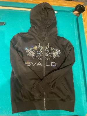BVALDI hoodie  - Svart hoodie med dragkedja och huva, dekorerad med ett glittrigt tryck av tre dobermans och texten 'BVALDI' på bröstet. Hoodien har långa ärmar och ribbade muddar vid ärmslut och nederkant. Perfekt för en cool och avslappnad stil.