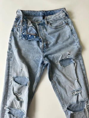 Ljusblå ripped jeans från Pull&Bear - Säljer ett par ljusblå jeans från Pull&Bear med flera slitningar och hål på benen. Jeansen har klassisk femficksdesign, bälteshällor och dragkedjegylf. Materialet är 100% bomull för en skön och avslappnad känsla.
