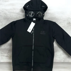 Svart hoodie med glasögon C.P. Company - Svart hoodie från C.P. Company med hel dragkedja, känguruficka och ribbade muddar. Huvan har inbyggda glasögon och metallöglor för en unik och cool look. Perfekt för dig som gillar streetwear och vill sticka ut i mängden.