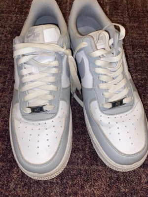 Nike Air Force 1 vita och grå sneakers - Nike Air Force 1 sneakers i vitt och ljusgrått läder med klassisk låg siluett. Skorna har perforerad tå, vita skosnören och den ikoniska Swoosh-loggan på sidan. Perfekta för dig som gillar stilrena och tidlösa sneakers.