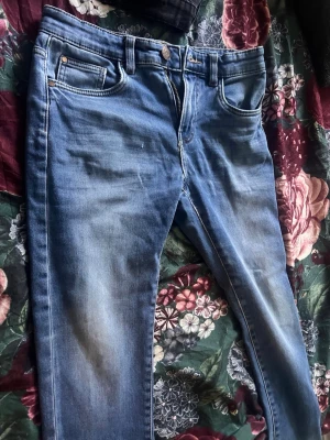 Blå jeans med slitningar - Ett par klassiska blå jeans med lätt slitna detaljer och fem fickor. Jeansen har normal midja, dragkedja och knappstängning. Materialet är denim i bomull och passformen är rak med något smalare ben.