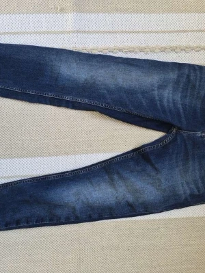 Blå skinny jeans från Tommy Jeans - Säljer ett par blå skinny jeans från Tommy Jeans med snygga slitningar och tvättade detaljer. Klassisk femficksmodell med gylf och knapp, samt tydliga sömmar i kontrastfärg. Perfekta för en avslappnad och trendig look.