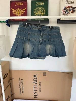 Minikjol i denim  - Snygg kort jeanskjol i mellanblå denim med utsvängd, veckad modell. Kjolen har flera djupa veck som ger den en fin A-linje och ett lite fylligare fall.  Klassisk jeansdetaljer med bälteshällor, knapp- och dragkedjestängning samt dekorativa sömmar/fickdetaljer framtill. Baktill är den ren och enkel med veckningen som tydlig detalj.  Fin att bära både till en söt/romantisk outfit, oversized topp eller mer Y2K/retro-inspirerad stil.