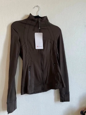 Brun träningsjacka från Lululemon - Slimmad brun träningsjacka från Lululemon med hög krage och hel dragkedja framtill. Jackan har långa ärmar, två fickor med dragkedja och är tillverkad i ett stretchigt syntetmaterial som passar perfekt till träning.