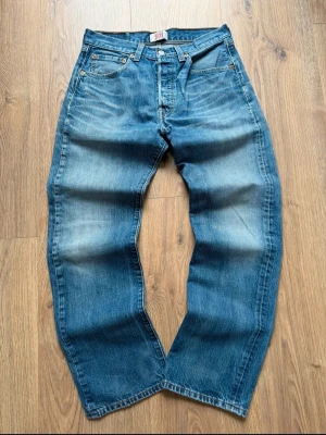 Levi's 501 blå raka jeans - Klassiska Levi's 501 jeans i mellanblå tvätt med raka ben och fem fickor. Jeansen har knappgylf och tydliga slitningar för en avslappnad look. Tillverkade i 100% bomull och har den ikoniska läderpatchen bak i midjan.