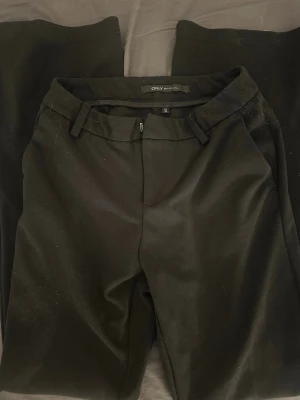 Svarta kostymbyxor från ONLY - Snygga svarta kostymbyxor från ONLY med klassisk rak passform och bälteshällor. Byxorna har en stilren look och stängs med dragkedja och hake. Perfekta för en clean och trendig outfit.