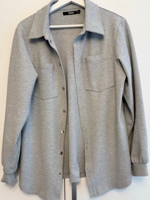 Oanvänd skjortjacka - Skön och snygg gråmelerad skjortjacka (overshirt) från Bik Bok i mjukt trikåmaterial. Har bröstfickor, krage, tryckknappar framtill samt manschetter vid ärmsluten. Perfekt att bära öppen över en topp eller stängd som skjorta.