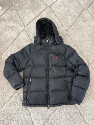 Svart Ralph Lauren dunjacka - .Säljer en riktigt snygg och varm dunjacka från Ralph Lauren i klassiskt svart utförande med den ikoniskt röda broderade loggan på bröstet. Perfekt för höst och vinter!  Märke: Polo Ralph Lauren  Färg: Svart med röd logga  Detaljer: Avtagbar/justerbar huva, bröstficka med dragkedja samt två sidfickor  Storlek: (Fyll i storlek här, t.ex. S/M/L eller juniorstorlek)  Skick: Very good condition / sparsamt använd utan defekter (justera om det finns något slitage) skriv om ni vill ha mått! 
