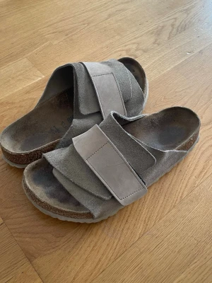 Birkenstock Kyoto - Säljer ett par grå sandaler i mocka med två breda kardborreband över foten. Sandalerna har en ergonomisk fotbädd och vit sula. Perfekta för chill dagar och enkel att ta på och av.