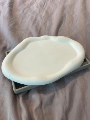Vit dekorativ skål i oregelbunden form - Säljer en vit dekorativ skål med oregelbunden, organisk form. Skålen är tillverkad i keramik och har en matt yta. Perfekt som inredningsdetalj eller för att förvara småsaker på skrivbordet eller i hallen. Köpte denna för att ha smycken på vilket har varit väldigt fint! Rensar nu ut denna eftersom jag köpte en annan mindre platta.
