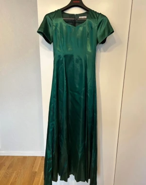 Grön långklänning i satin - Elegant grön långklänning i glansigt satinliknande material med korta ärmar och rund halsringning. Klänningen har en lång dragkedja längs ryggen, och ett klassiskt, tidlöst snitt som ger en lyxig känsla. Finns i Vallberga, Laholms kommun 