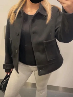 Svart ulljacka med stora fickor - Stilren svart jacka i ull med stora framfickor och knappar framtill. Jackan har en klassisk krage och långa ärmar med dekorativa detaljer vid ärmsluten. Passar perfekt till både jeans och kjol för en trendig look.