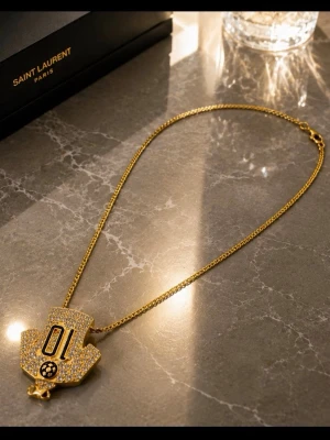 Guldigt halsband med stor bling-berlock - Unikt halsband i guld med en stor, glittrande berlock täckt av små stenar och siffran 01 i svart. Berlocken har en cool, geometrisk form och en liten detalj längst ner. Kedjan är tunn och elegant, perfekt för att sticka ut.