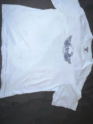 Vit t-shirt med tryck från Affliction - Säljer en vit t-shirt från Affliction med stort grafiskt tryck i svart på både fram- och baksidan. T-shirten har klassisk passform och korta ärmar. Perfekt för dig som gillar streetwear och vill sticka ut med cool design.