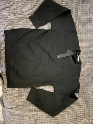 Svart sweatshirt från Stone Island - Säljer en svart sweatshirt från Stone Island med den klassiska loggan på vänster ärm. Tröjan har rund halsringning, ribbade muddar och är gjord i mjukt bomullsmaterial. Perfekt för dig som gillar streetwear och vill ha en clean look.