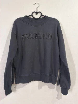 Mörkgrå sweatshirt med STOCKHOLM-tryck - Cool mörkgrå sweatshirt med texten STOCKHOLM broderat framtill. Tröjan har rund hals, långa ärmar och dragkedjor i sidorna för extra stil. Tillverkad i mjukt bomullsmaterial och har ribbade muddar vid ärmslut och nederkant. Perfekt för en avslappnad streetwear-look.