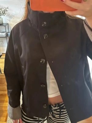 Svart jacka med hög krage och knappar - Snygg svart jacka med hög krage och stora knappar framtill. Jackan har långa ärmar med kontrasterande beige muddar och en rak passform. Perfekt för dig som gillar stilrena och minimalistiska plagg.