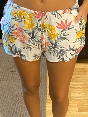 Blommiga shorts med dragsko - Snygga och bekväma shorts med blommigt mönster i rosa, gul och grön på vit bakgrund. De har en praktisk dragsko i midjan för justerbar passform. Storlek 38/M, men passar även mig som brukar ha 34-36/xs-s!